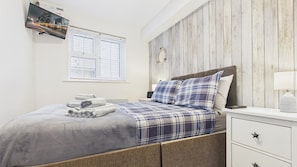 A bedroom with a bed, a nightstand, a lamp, a wall-mounted TV, and a window with blinds.