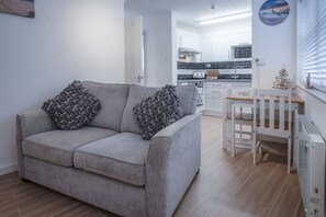 A living room with a grey sofa and two decorative pillows, a wooden dining table with a white chair, and a kitchen in the background. - Castle Cove - 1 Bedroom Apartment - Tenby (Tenby)