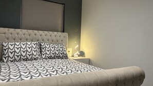 Camera da letto con testiera trapuntata, cuscini decorati, lampada da comodino e colori neutri.