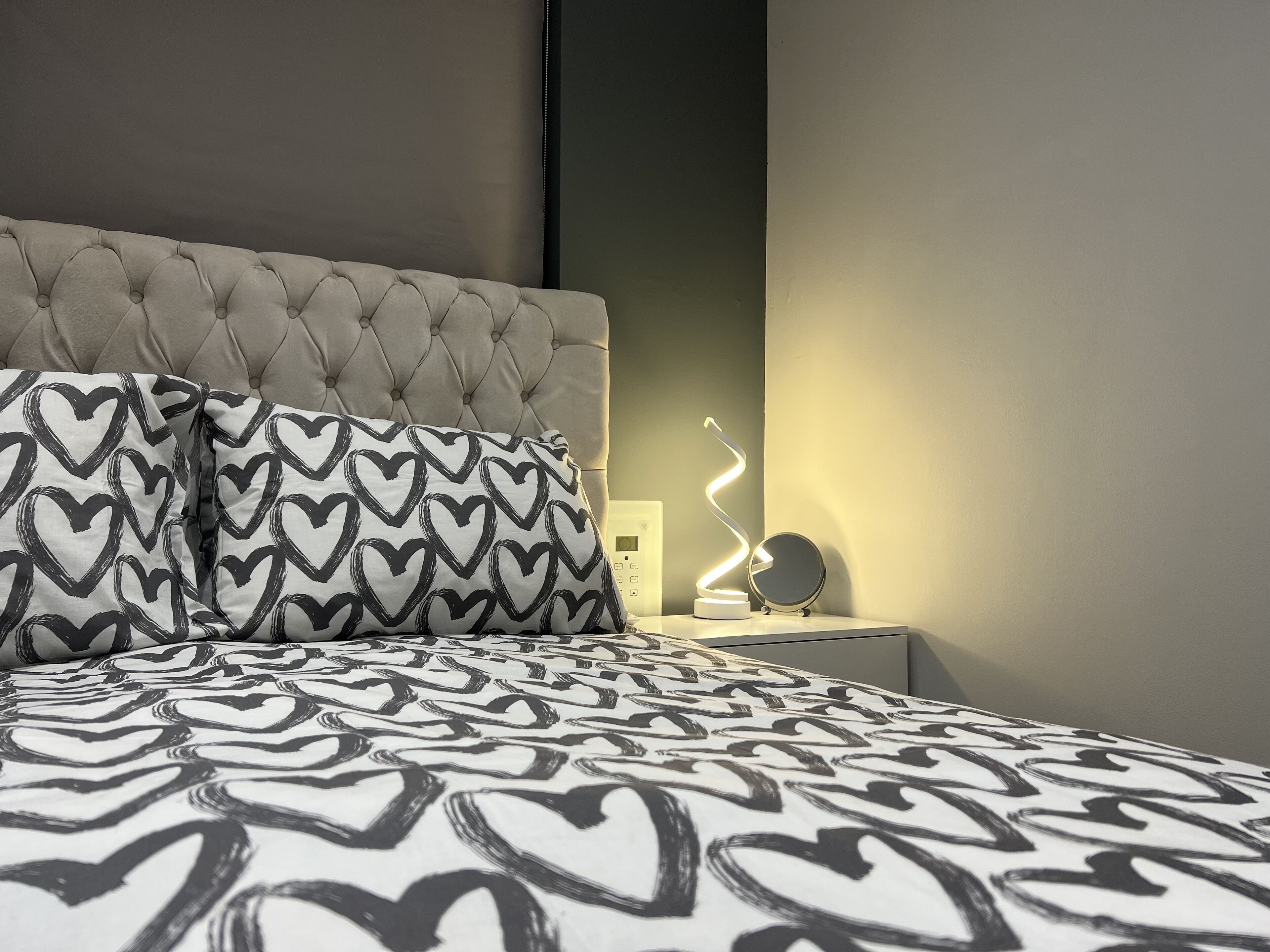 Quarto com cabeceira estofada, almofadas com padrão de coração e mesa de cabeceira com espelho e luminária.