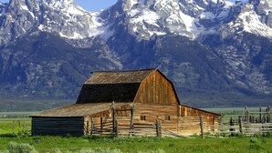 Un granero de madera rústico con techo inclinado, rodeado por una cerca y con montañas nevadas al fondo.
