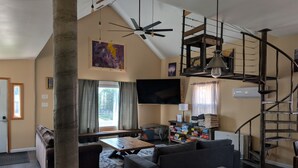 A spacious living area with a high ceiling, a modern staircase, a flat-screen TV, and a large window with curtains.