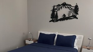 A neatly made bed with a white headboard and dark blue bedding, a small bedside table with a lamp, and a wall decoration of a mountain scene. - Bright Condo With Views & Easy Access to CDA River.  Minutes From Gondola! (Kellogg)