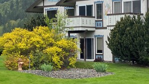 A two-story building with a balcony, surrounded by a well-maintained lawn and a mountain backdrop. - Bright Condo With Views & Easy Access to CDA River.  Minutes From Gondola! (Kellogg)