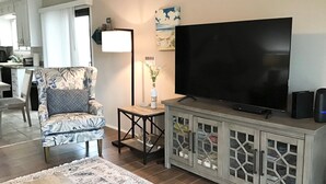 Ein Wohnzimmer mit einem großen Flachbildfernseher, einem gemusterten Sessel, einem kleinen Beistelltisch mit einer Lampe und einer Vitrine.