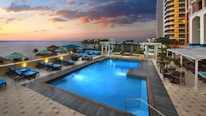 A pool area with lounge chairs and umbrellas, a building with balconies, and a view of the ocean at sunset. - Marriotts BeachPlace Towers - 2 Bedroom (Fort Lauderdale)