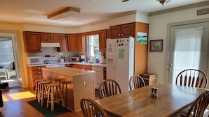Cozinha com armários de madeira, ilha central com banquetas, mesa de jantar com cadeiras e geladeira com imãs.