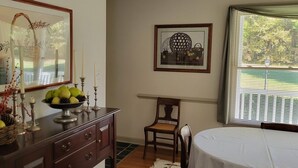Quarto com mesa de jantar, uma cadeira de madeira, cômoda com uma fruteira, espelho emoldurado e janela com persianas.