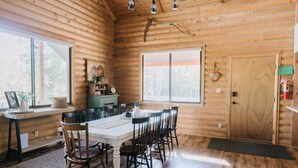 Interior de una cabaña de madera con una mesa de comedor, sillas y un ventanal grande.