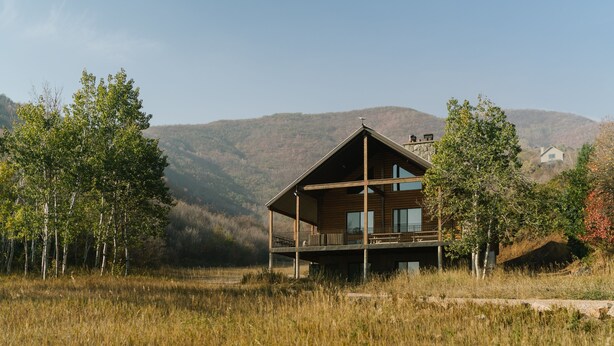 Una cabaña de madera moderna con balcón, rodeada de hierba alta y árboles, con montañas al fondo.