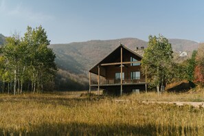 Una cabaña de madera moderna con balcón, rodeada de hierba alta y árboles, con montañas al fondo.