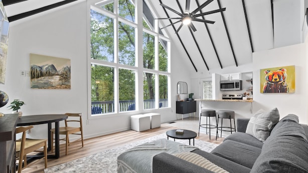 A modern living room with a gray sofa, wooden dining table, and chairs. There is a kitchen area with a microwave and a bar stool. Large windows provide natural light and a view of trees. - Escape to Shenandoah River, Hot Tub after a Hike!  (Front Royal)