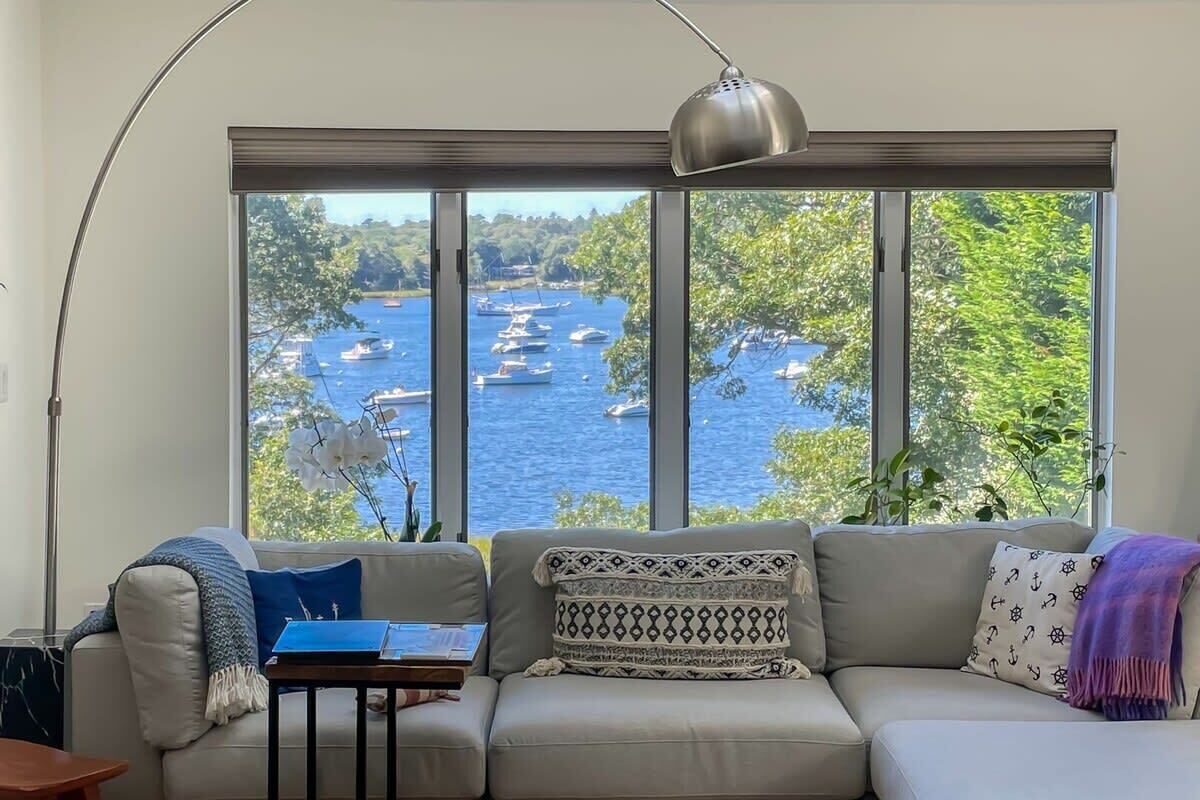 Sala de estar com uma grande janela com vista para um corpo de água com barcos, um sofá, uma mesa com um livro e uma luminária.