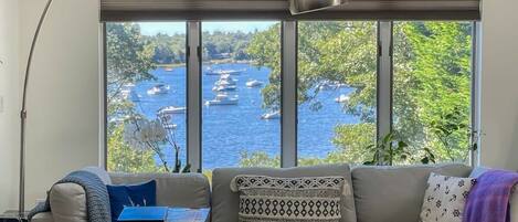 A living room with a large window overlooking a body of water with boats, a sofa, a table with a book, and a lamp.