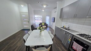Eine moderne Küche mit grauen Schränken, einer weißen Arbeitsplatte und Edelstahlgeräten. Es gibt eine Essecke mit einem weißen Tisch und Stühlen, und ein Sofa im Hintergrund.