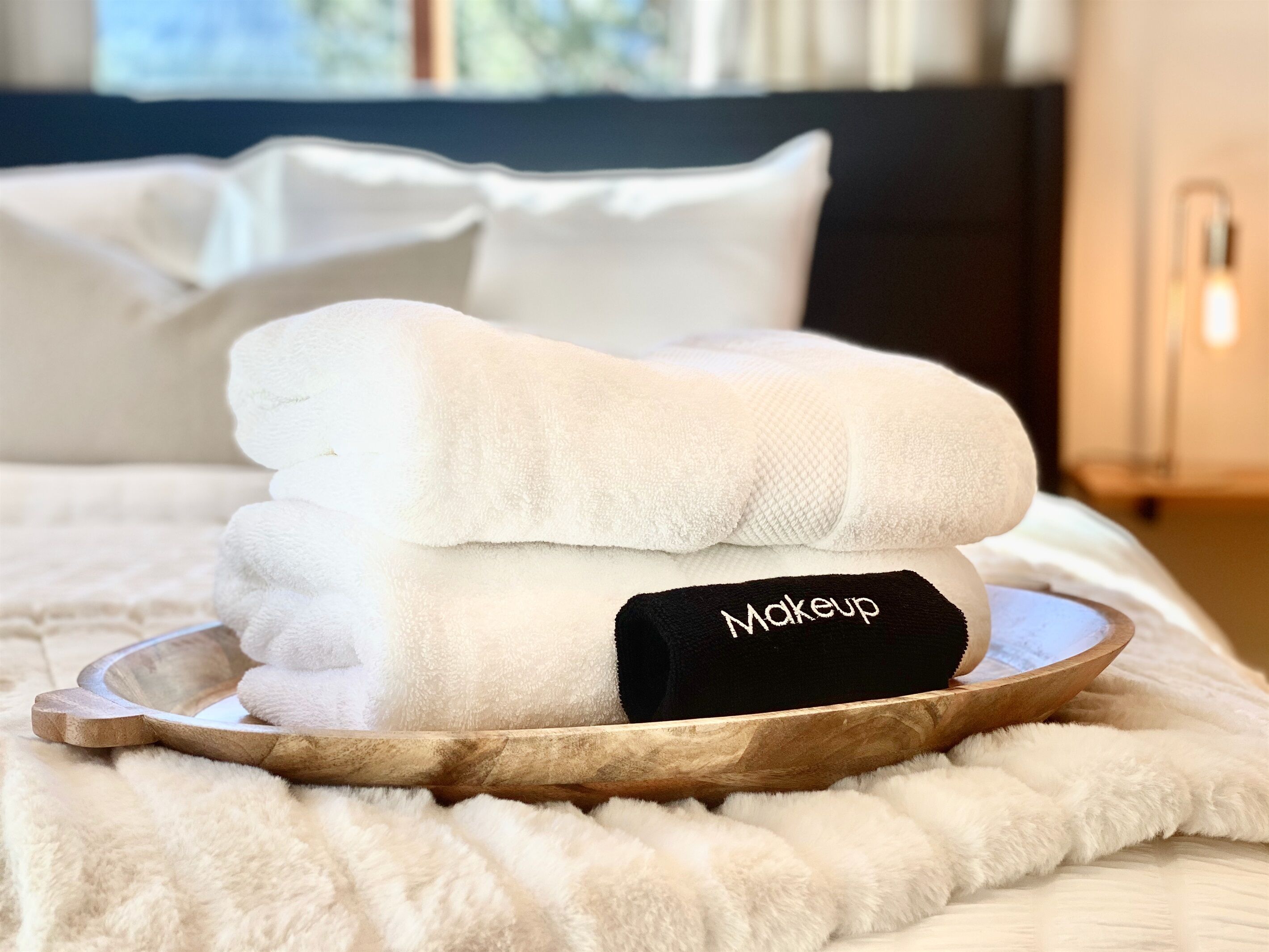 Una toalla blanca doblada con una etiqueta negra que dice 'Maquillaje', sobre una bandeja de madera, colocada en una cama con ropa blanca.