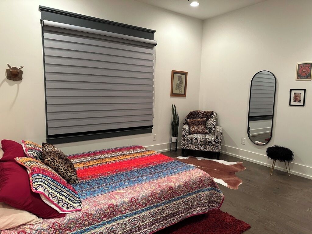 A bedroom with a large bed, a chair, a mirror, and a window with blinds.