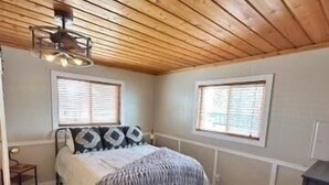 Ein Schlafzimmer mit Holzboden, einem Bett mit gemusterten Betttextilien, zwei Fenstern mit Jalousien und einem kleinen Tisch.
