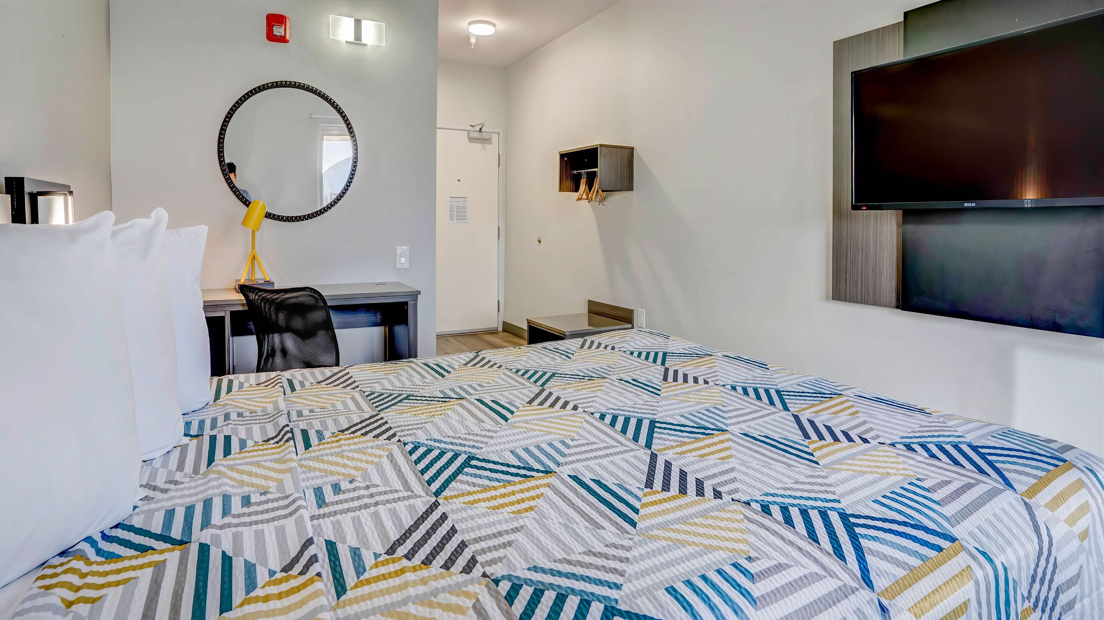 Une chambre d’hôtel avec un grand lit, un bureau avec une chaise, un miroir rond, une télévision et un petit meuble.
