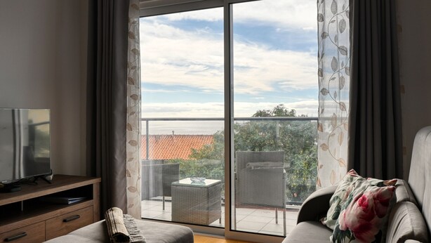 A modern living room with a sofa, a television on a wooden stand, and a balcony with a table and chairs. - Tamariz Sea View, a Home in Madeira (Caniço)