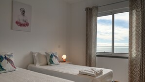 A hotel room with two beds, a window with a view, a decorative pillow, and a wall-mounted artwork. - Tamariz Sea View, a Home in Madeira (Caniço)