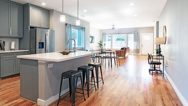 A modern kitchen with grey cabinets, a central island with stools, and a dining area with a table and chairs. - 3BR Comfy Bungalow 1747! Medical Center, Stadiums! ⭐️⭐️⭐️⭐️⭐️ (Houston)
