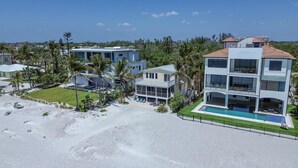 Une propriété en bord de mer, composée de plusieurs étages, dotée d’une piscine et entourée de palmiers et d’une plage de sable.