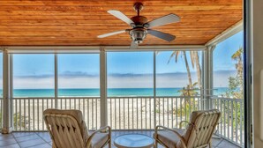 Un espace extérieur abrité, doté de deux fauteuils en osier et d’une petite table, offrant une vue sur une plage bordée de palmiers et une vue dégagée sur l’océan.