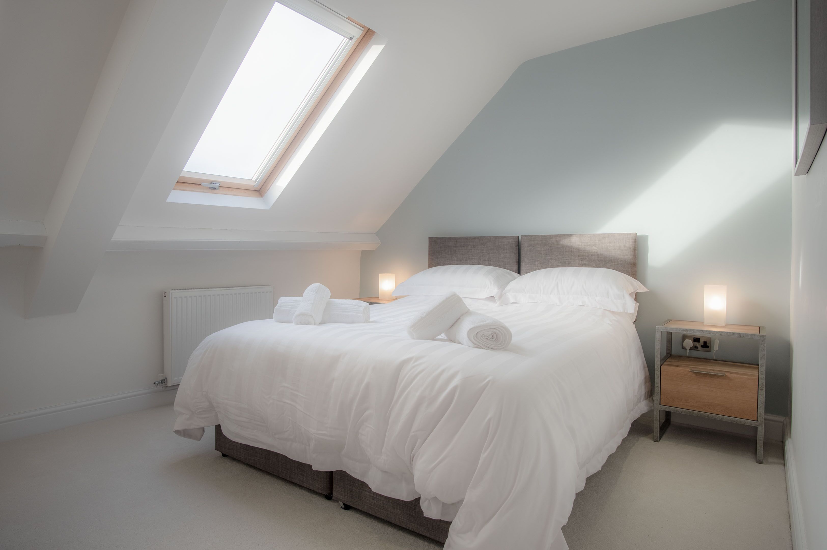 A bedroom with a skylight, a bed with white linens, a bedside table with a lamp, and a radiator.
