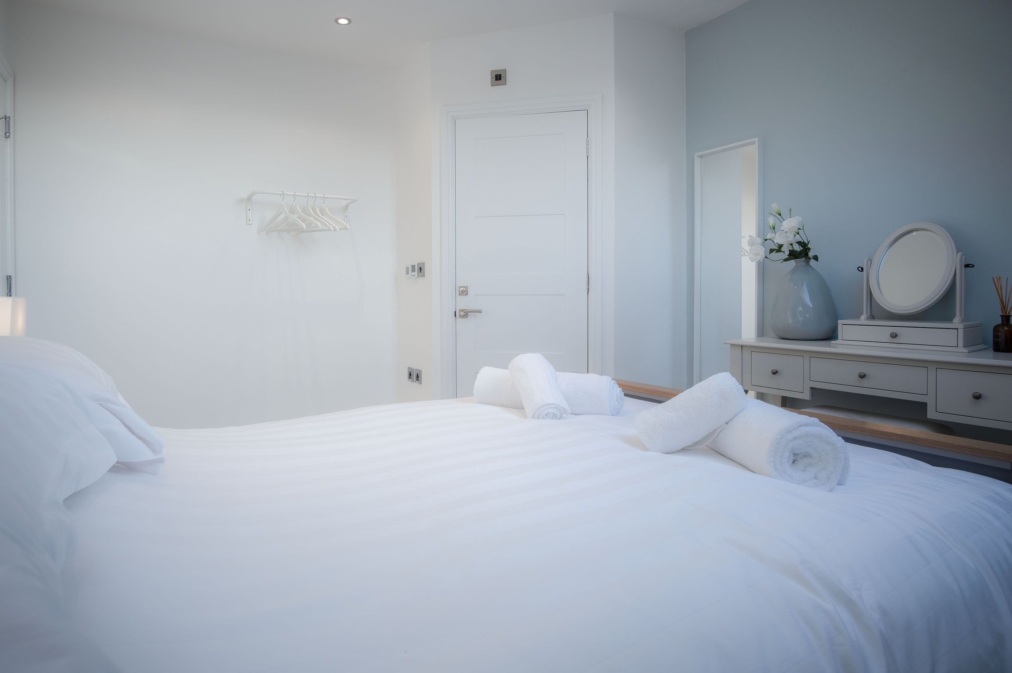 Una cama bien hecha con sábanas blancas y toallas enrolladas, una puerta blanca, un tocador con espejo y un jarrón con flores.