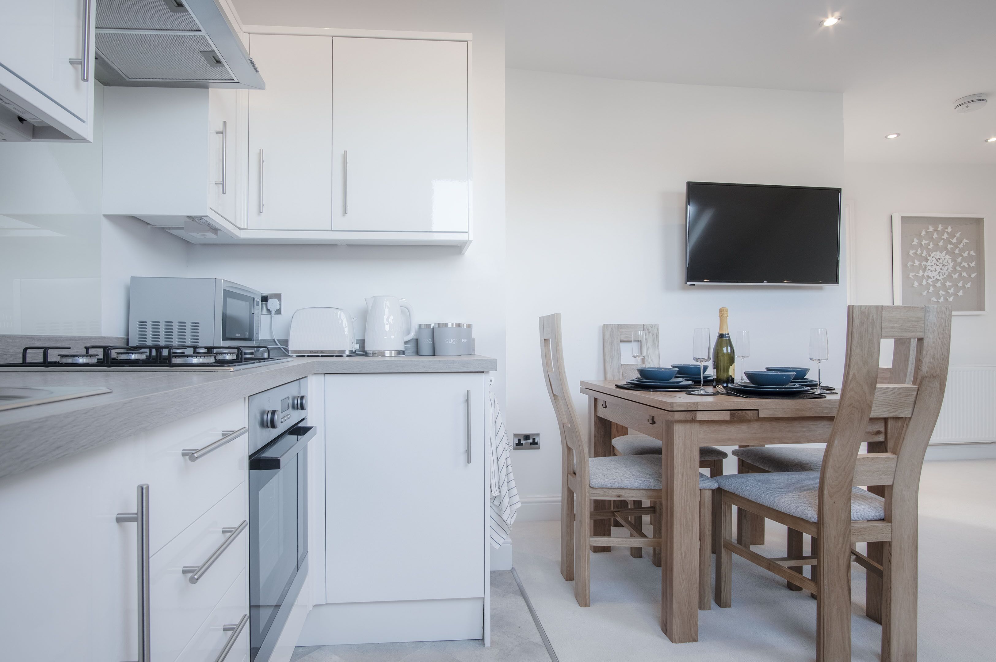 A modern kitchen with white cabinetry, a built-in oven, a microwave, and a gas stove. There is a dining area with a wooden table and chairs, and a flat-screen TV mounted on the wall.