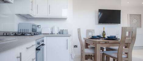 A modern kitchen with white cabinetry, a built-in oven, a microwave, and a gas stove. There is a dining area with a wooden table and chairs, and a flat-screen TV mounted on the wall.