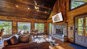 A spacious living room with a stone fireplace, leather sofas, and large windows offering a view of the outdoors. - Fireplaces, hot tub, romantic cabin perfect for a honeymoon or a getaway for two (Ringold)