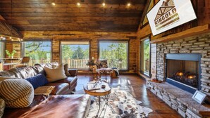 A rustic cabin interior with a stone fireplace, wooden floors, and a large window overlooking greenery. - Fireplaces, hot tub, romantic cabin perfect for a honeymoon or a getaway for two (Ringold)