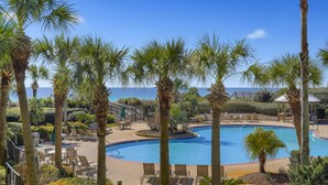A pool area with palm trees, lounge chairs, and a view of the ocean. - The Crescent unit 110 2BR Condo w/ FREE Beach Service! (Miramar Beach)