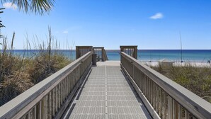 A wooden boardwalk leading to a beach with clear blue water and a palm tree. - The Crescent unit 110 2BR Condo w/ FREE Beach Service! (Miramar Beach)