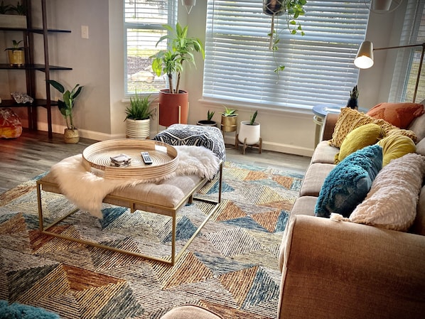A cozy living room with a sofa, a coffee table, and a patterned rug. There are several potted plants and a window with blinds.