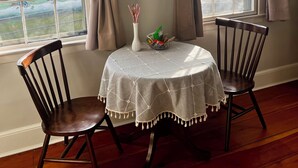 Een eethoek met een ronde tafel bedekt met een witte tafelkleed, twee houten stoelen, een vaas met bloemen en een lijst met een afbeelding aan de muur.
