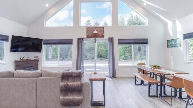 En rummelig stue med højt til loftet, store vinduer, pejs og spiseområde med bænke.
