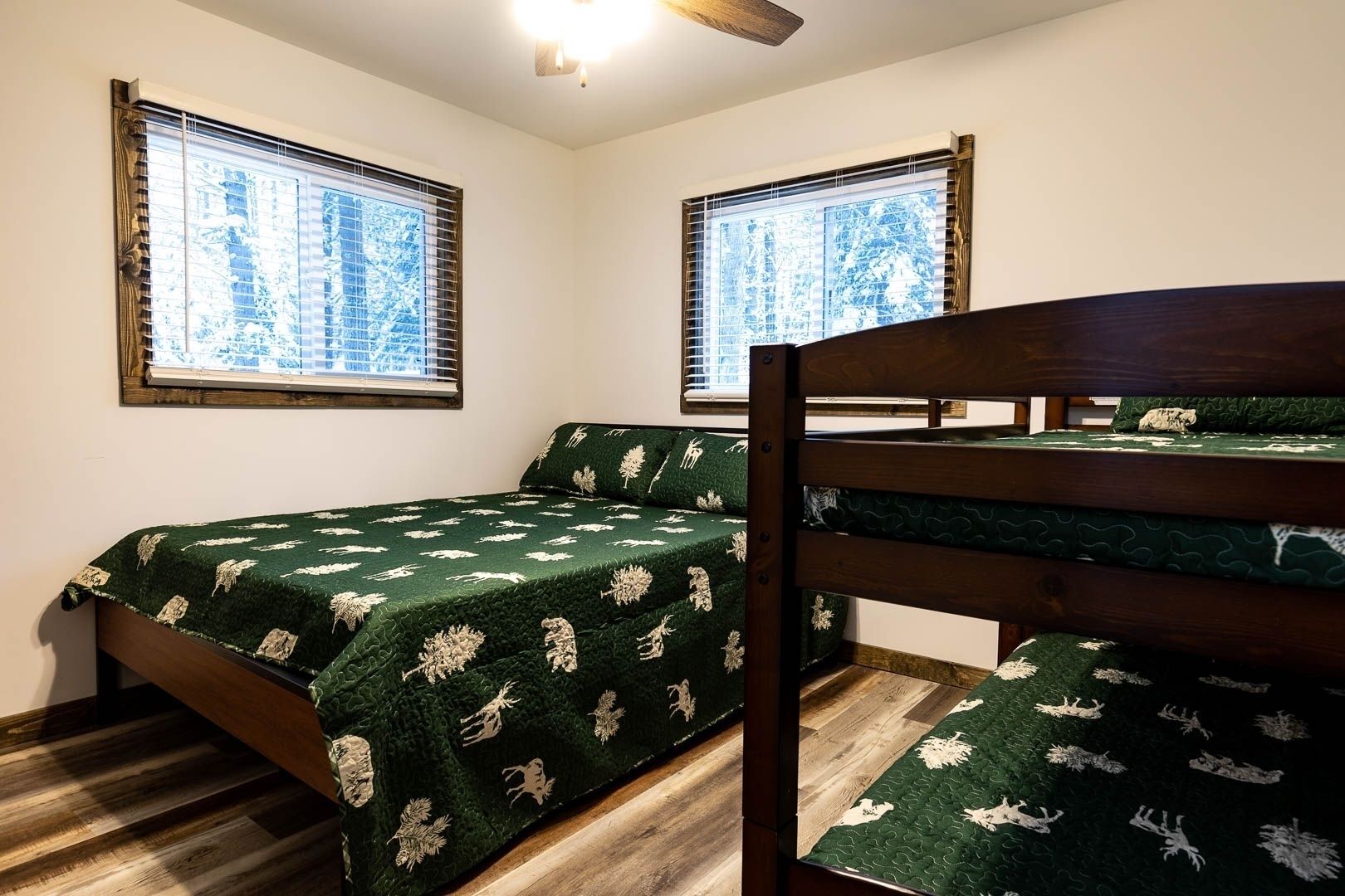 A bedroom with two beds, one with a wooden headboard and the other with a metal frame. There are two windows with blinds, and the room has a wooden floor.