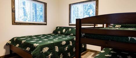 A bedroom with two beds, one with a wooden headboard and the other with a metal frame. There are two windows with blinds, and the room has a wooden floor.
