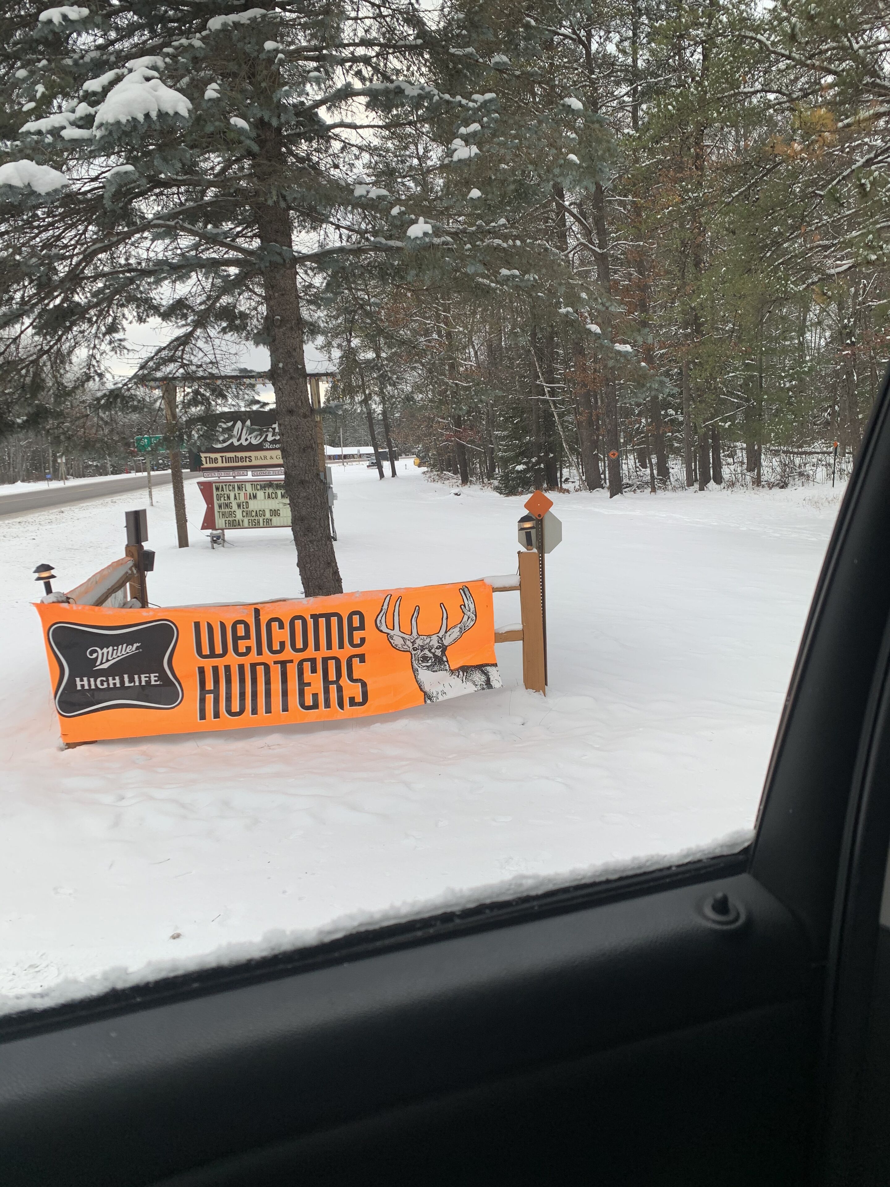 A snow-covered welcome sign for hunters with a deer illustration, a Miller High Life logo, and a sign for The Timbers Bar & Grill.