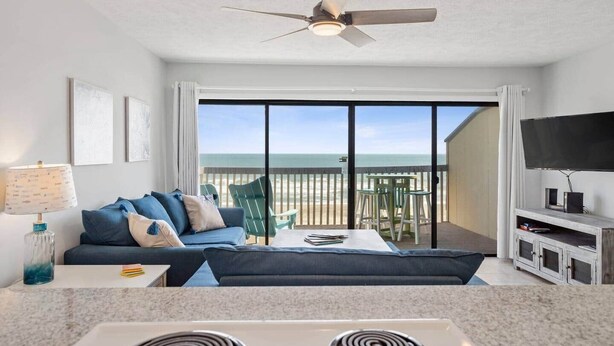 Una sala de estar con sofá, mesa de centro y televisor sobre un mueble bajo. Hay un balcón con vista a la playa.