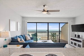 A living room with a sofa, coffee table, and a TV on a stand. There is a balcony with a view of the beach. - Updated beachfront condo with stunning views! (Carolina Beach)