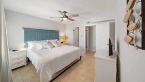 Un dormitorio con cama, mesitas de noche, ventilador de techo, televisor montado en la pared y una puerta que da a otra habitación.