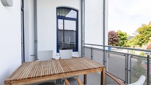 Ein Balkon mit Holztisch und Korbstühlen, ein Fenster mit blauer Umrandung und einem Metallgeländer.
