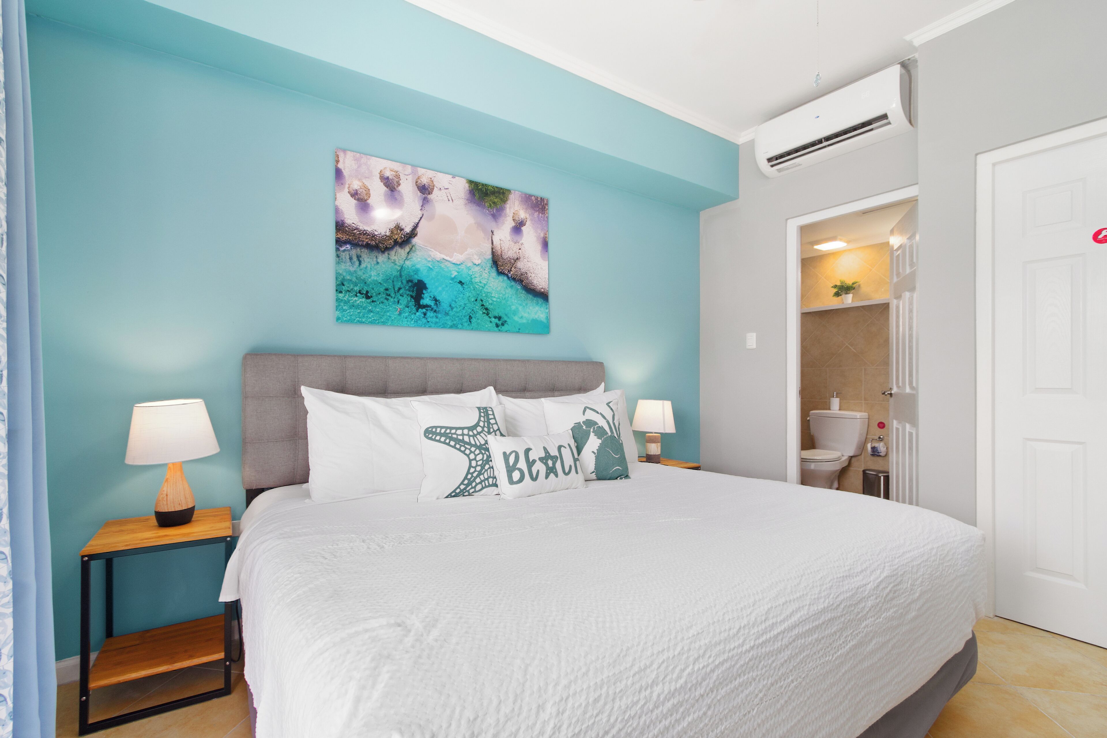 Une chambre à coucher avec un lit, deux tables de chevet avec des lampes, un tableau accroché au mur et une salle de bain visible à travers une porte ouverte.