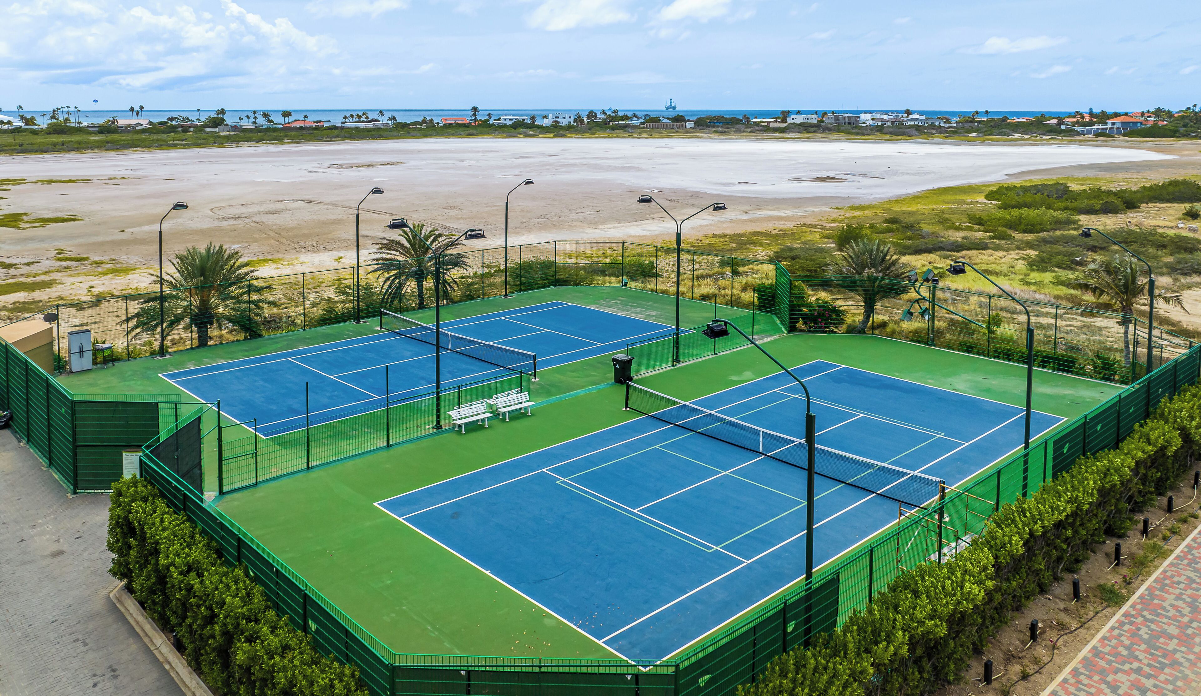 A tennis court complex with multiple courts, surrounded by greenery and a beach view.