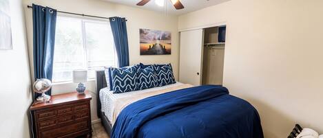 Een slaapkamer met een bed, een dressoir, een plafondventilator, een raam met gordijnen, een schilderij en een deur.