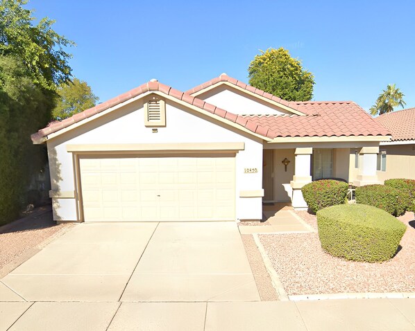 A single-story house with a beige garage door, a red-tiled roof, and a small front yard with a trimmed bush.